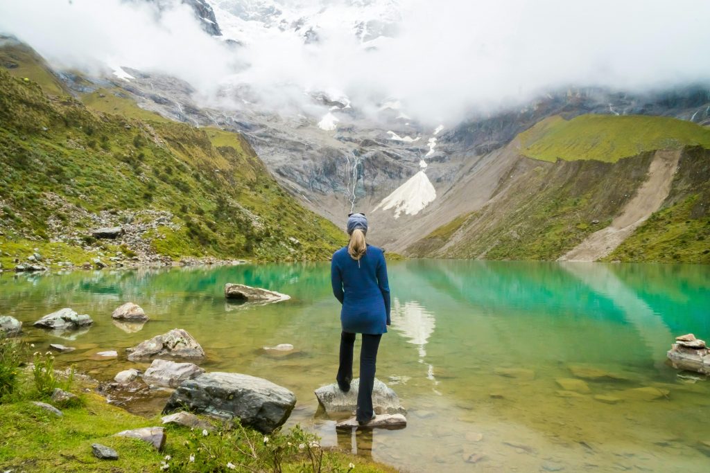 A woman stands beside a turquoise lake with fog-covered Andes mountains in the background.