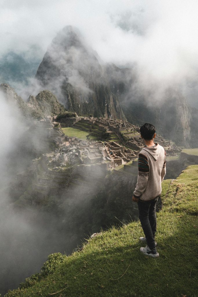 A man stands on a cliff edge overlooking the ancient ruins of Machu Picchu enveloped in mist.